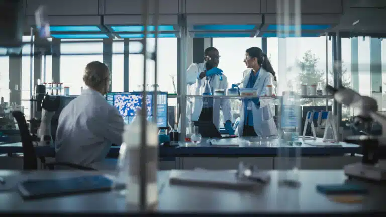 Scientists wearing lab coats and safety goggles working collaboratively in a modern laboratory with advanced equipment and computers in the background.